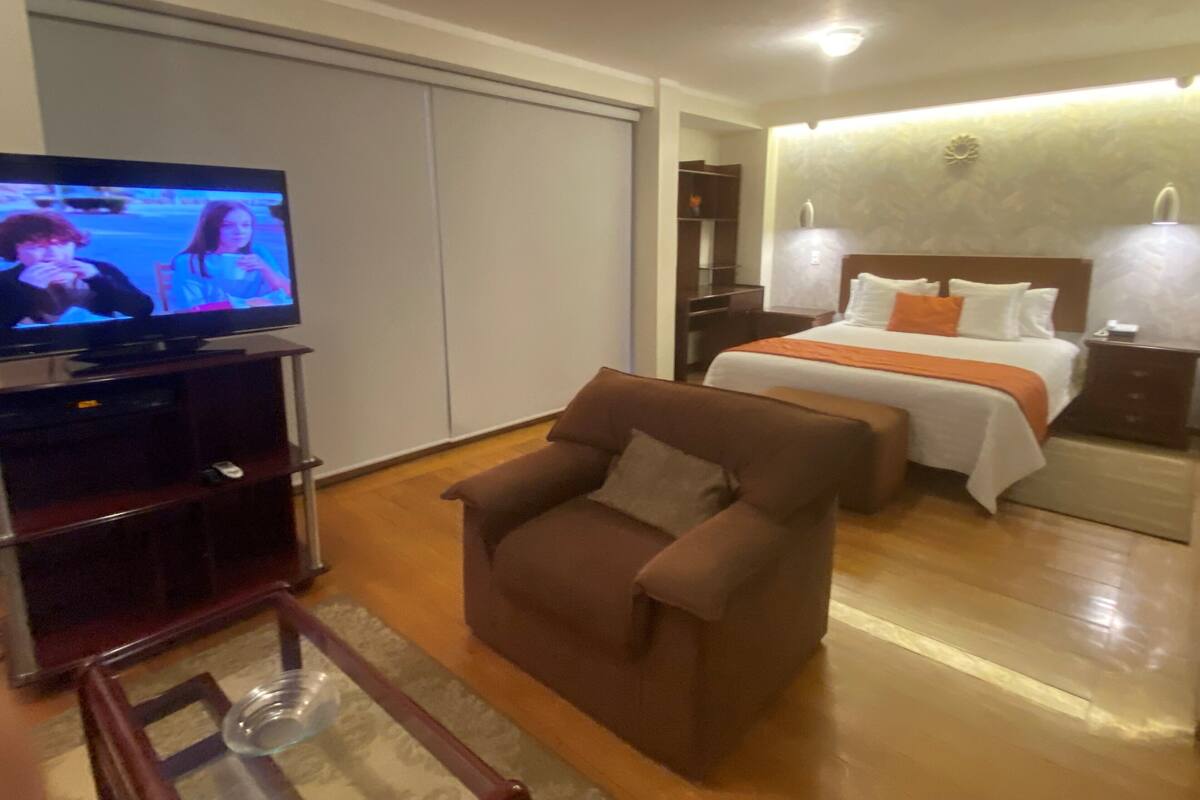A hotel room with a television, a bed with white and orange bedding, a brown armchair, and a wooden floor.