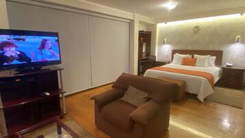 A hotel room with a television, a bed with white and orange bedding, a brown armchair, and a wooden floor.