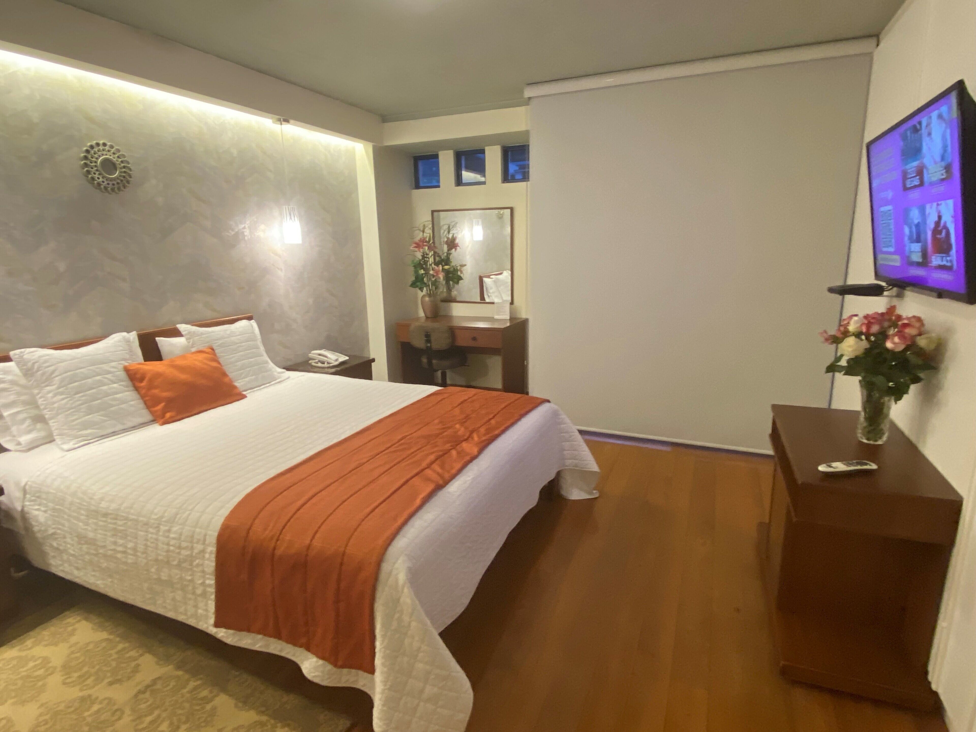 Habitación de hotel con cama, escritorio, silla, espejo, un florero con flores y televisor en la pared.