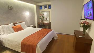 Habitación de hotel con cama, escritorio, silla, espejo, un florero con flores y televisor en la pared.