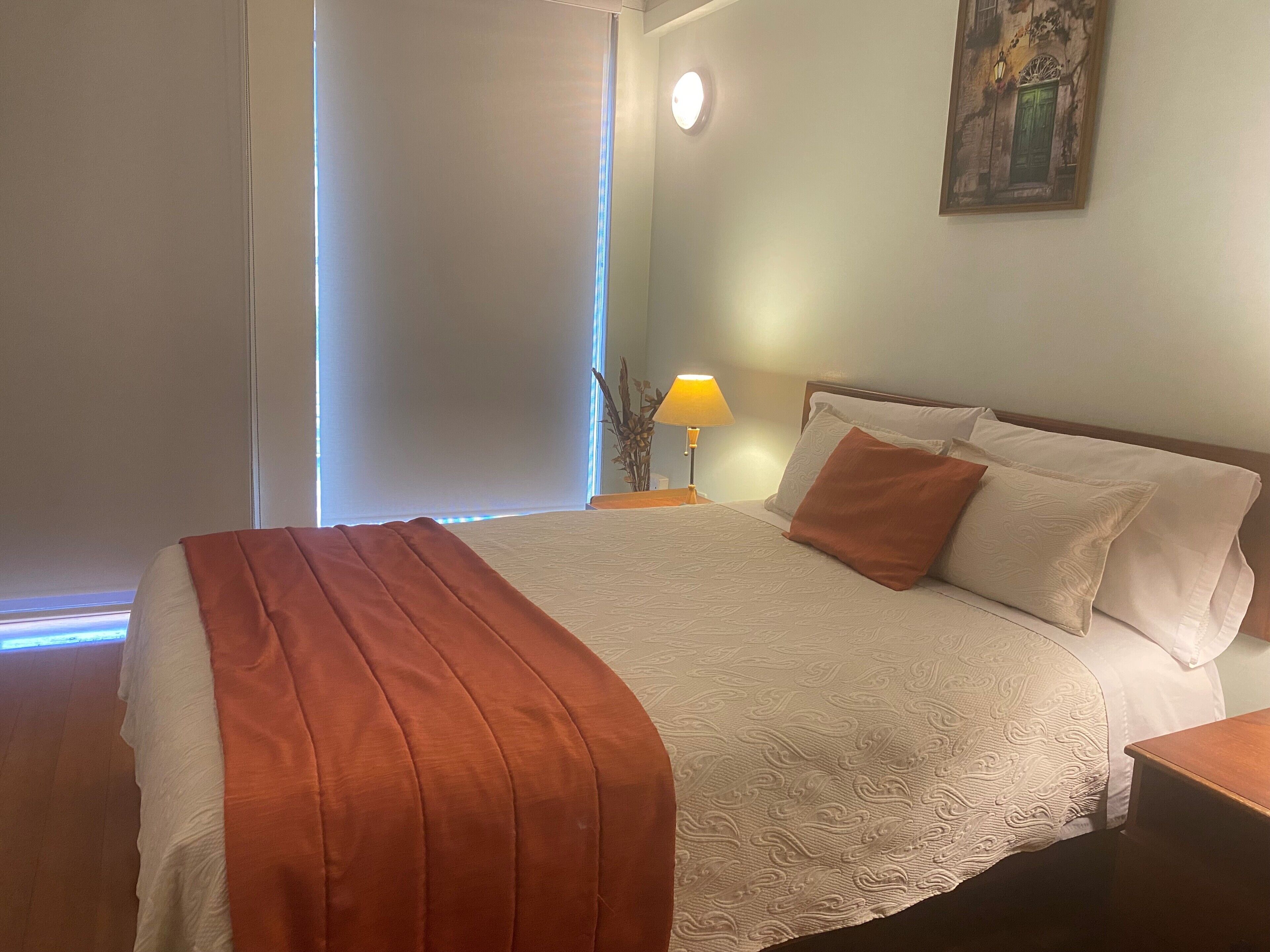 Ein ordentlich bezogenes Bett mit einer weißen und orangefarbenen Decke, einer Nachttischlampe und einem Bild an der Wand.