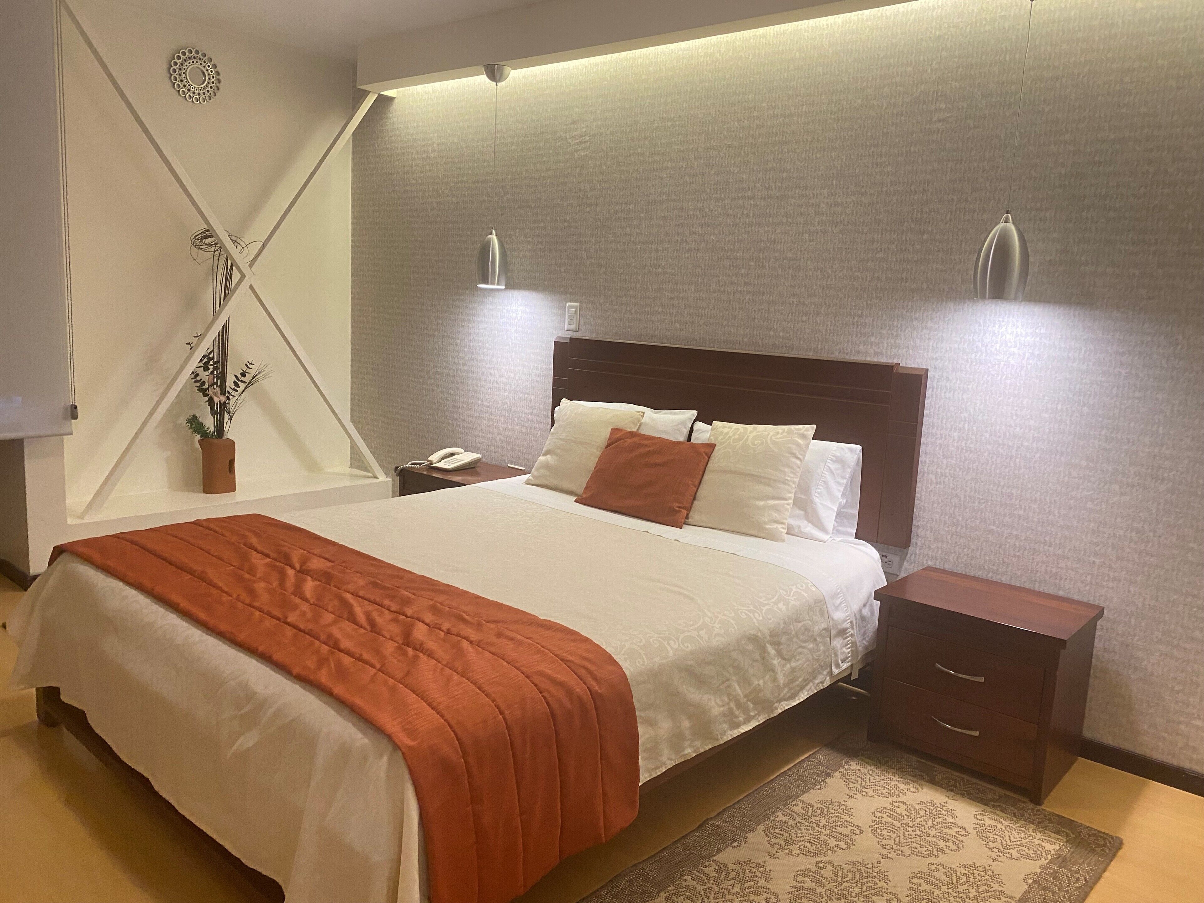 A neatly made bed with a white and orange bedspread, a bedside table with a phone, and a nightstand with a lamp.