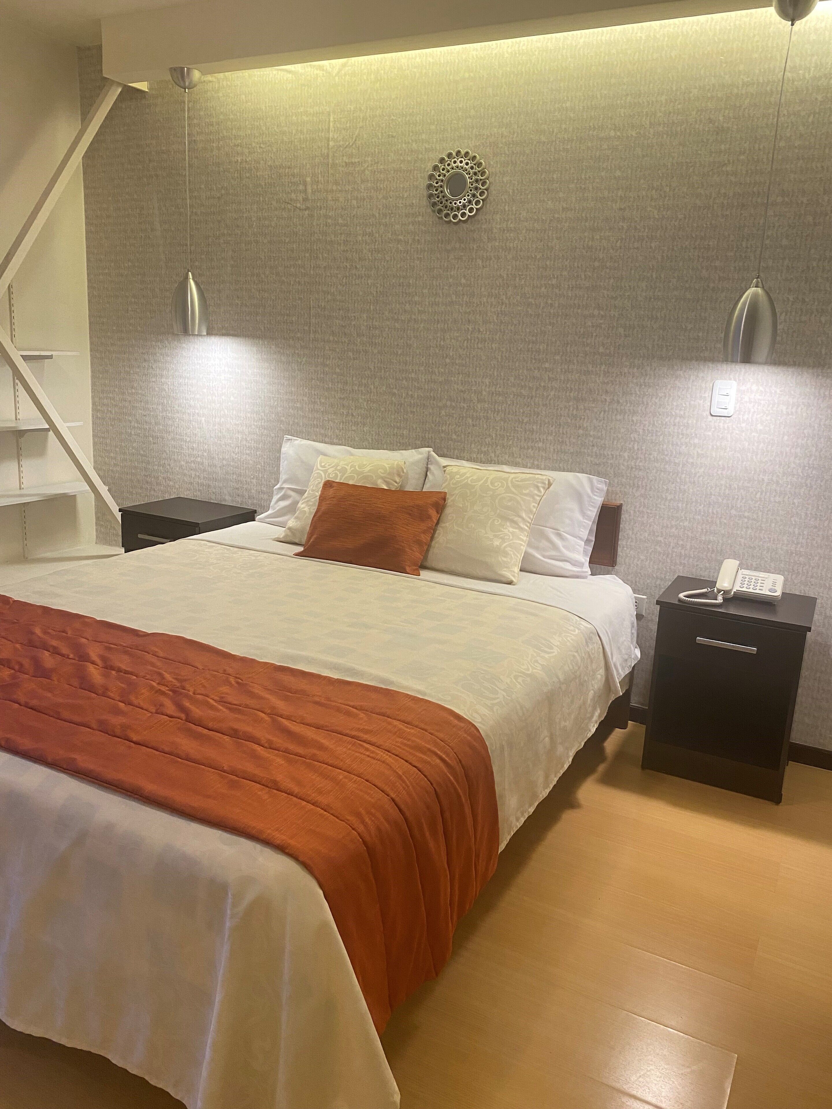 Una cama bien hecha con colcha blanca y naranja, una mesita de noche negra con un teléfono, y un aplique de pared.