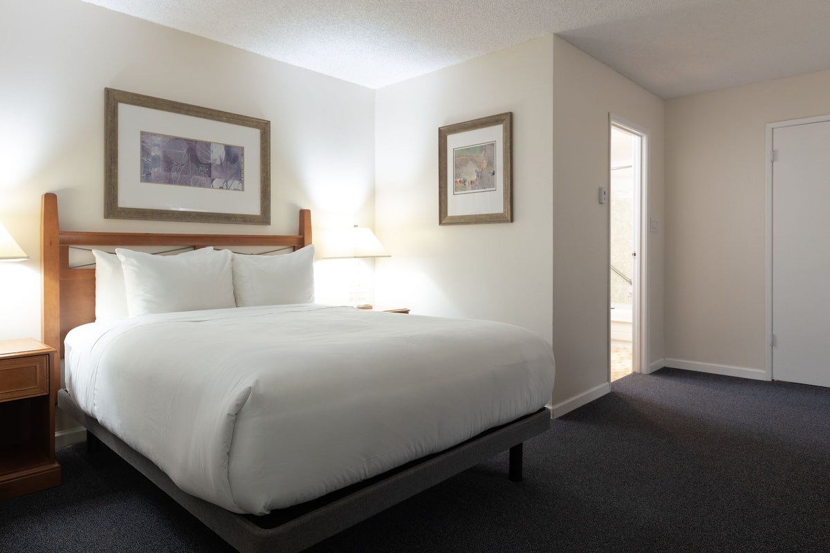 A neatly made bed in a hotel room with a wooden headboard, two framed pictures on the wall, a nightstand with a lamp, and a door leading to another room.