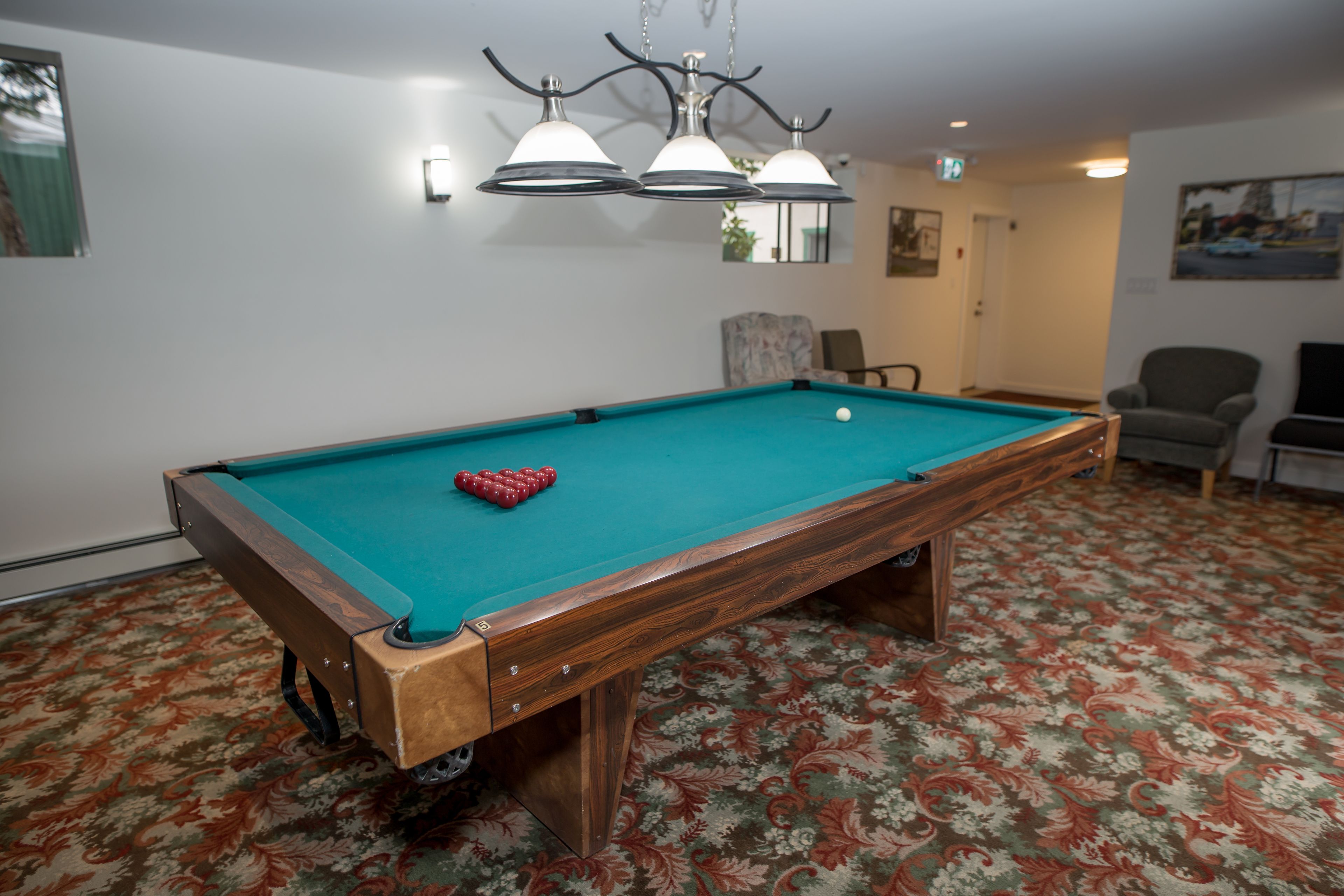 A pool table with a green felt surface, a red cue ball, and a pool cue. There are two wall-mounted lamps above the table. The room has patterned carpet flooring and a door in the background.