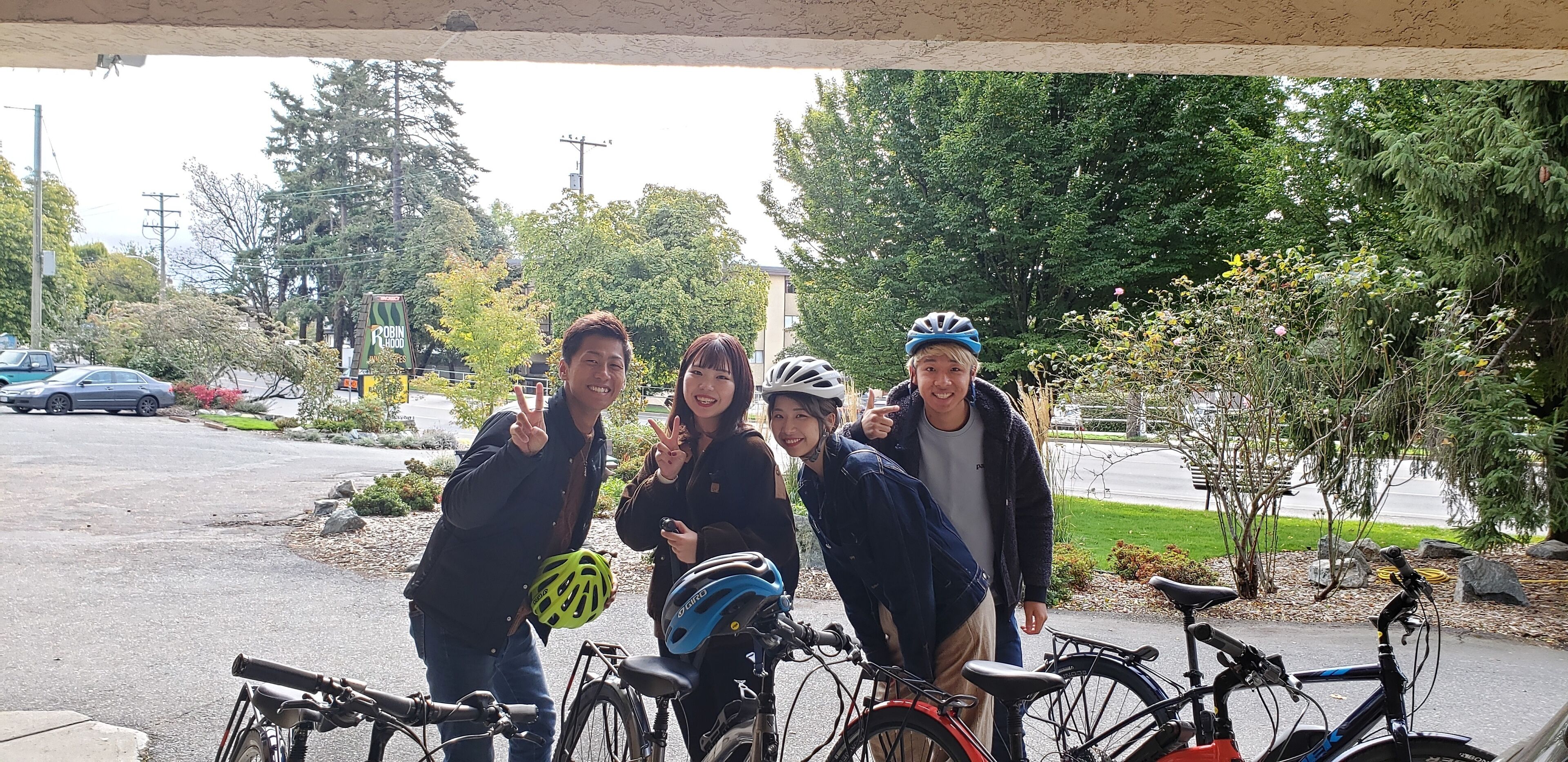 A group of people posing with bicycles in a covered outdoor area with trees and a road in the background.