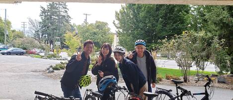 A group of people posing with bicycles in a covered outdoor area with trees and a road in the background.