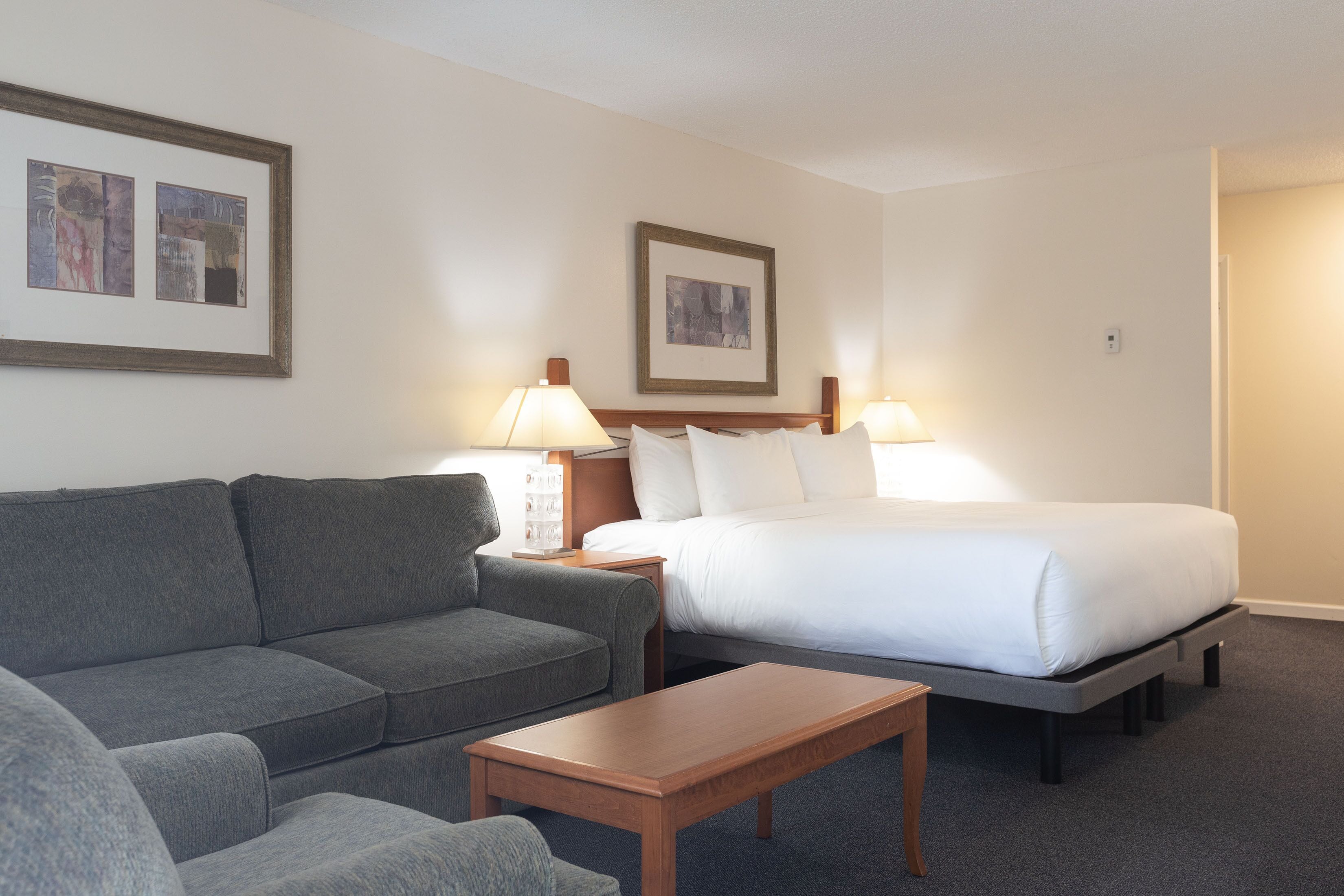 A hotel room with a grey sofa, a wooden coffee table, a bed with white linens, two bedside lamps, and two framed pictures on the wall.