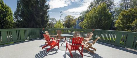 An outdoor patio with a round table and four Adirondack chairs, surrounded by a green fence and trees.