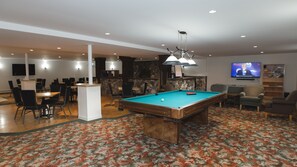 A pool table in a room with a patterned carpet, a television, and a seating area with chairs and a sofa.