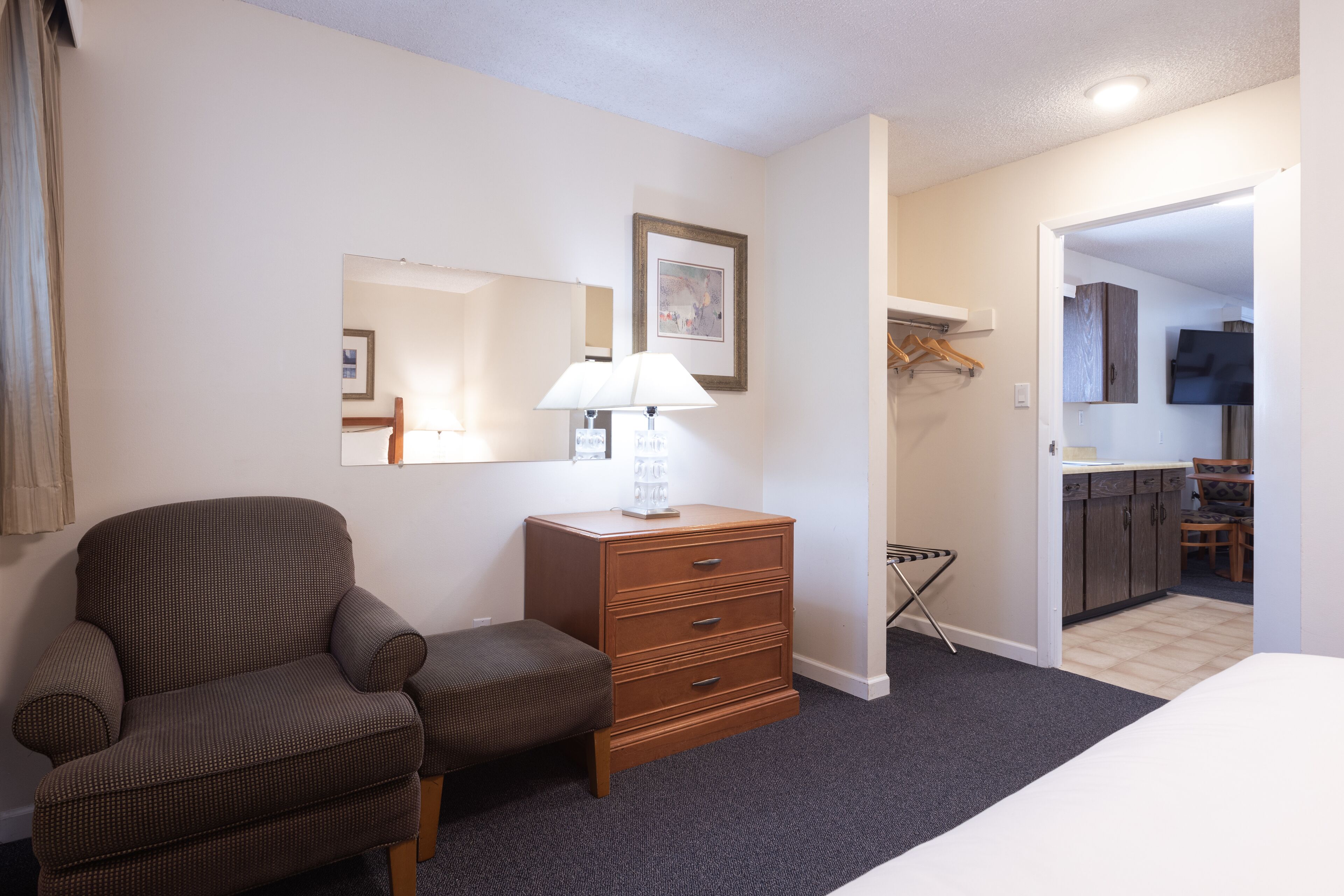 Une chambre d’hôtel avec un lit, une chaise, une commode, une lampe, un miroir et un coin cuisine visible à travers une porte ouverte.