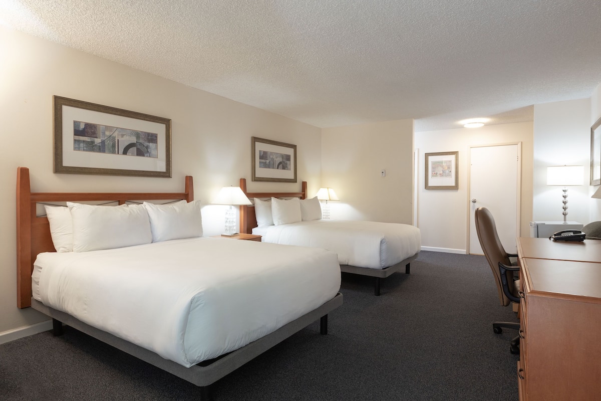 A hotel room with two beds, a desk, and a chair. There are framed pictures on the walls and a door in the background.