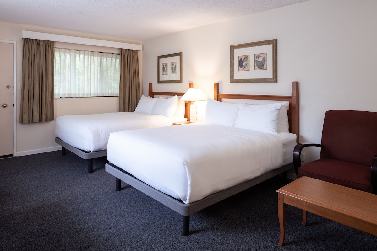A hotel room with two single beds, a wooden nightstand with a lamp, a wooden chair, and framed pictures on the wall.