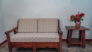 Un banco de madera con un cojín estampado, una pequeña mesa de madera con un florero de flores rojas, y un suelo de baldosas de piedra.