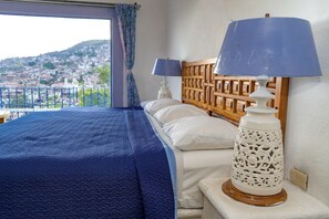 A bedroom with a view of a cityscape, a wooden headboard, two bedside lamps, and a blue bedspread. - Hotel de la Borda (Taxco)