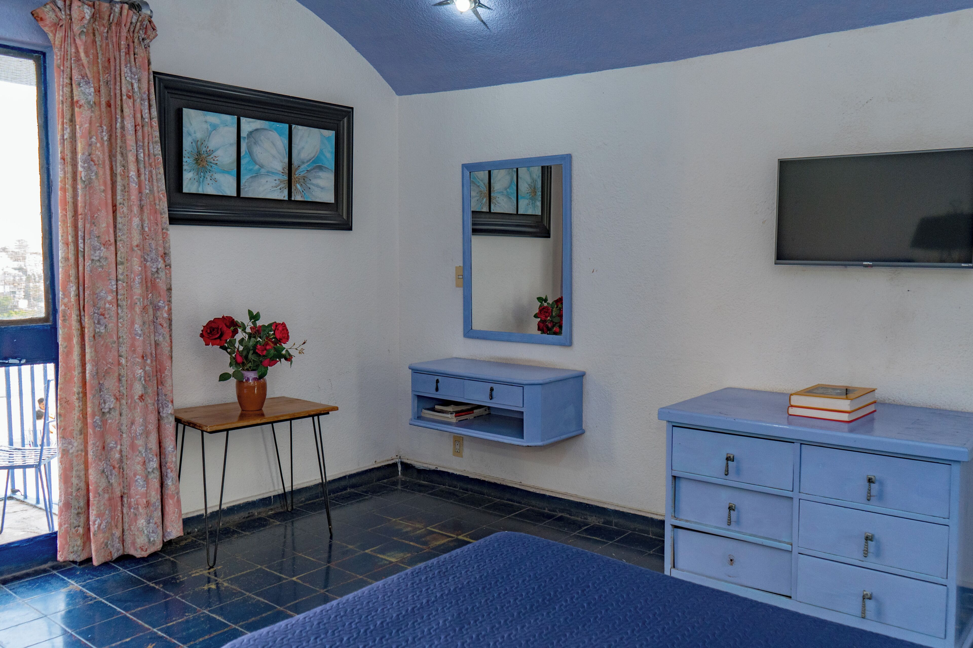 A room with a bed, a small table with a vase of flowers, a blue dresser, a mirror, and a TV mounted on the wall.