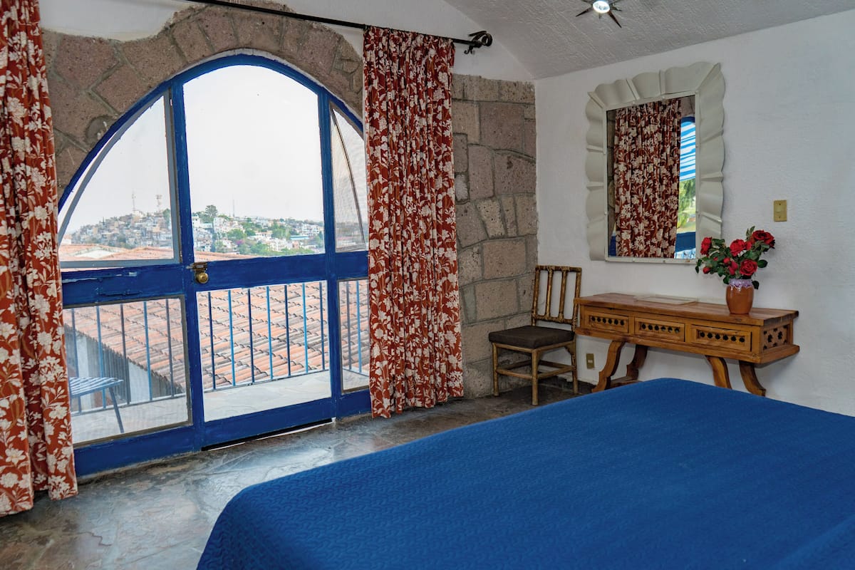 Una habitación con un amplio ventanal arqueado, una cama, un aparador de madera, una silla y un jarrón de flores.