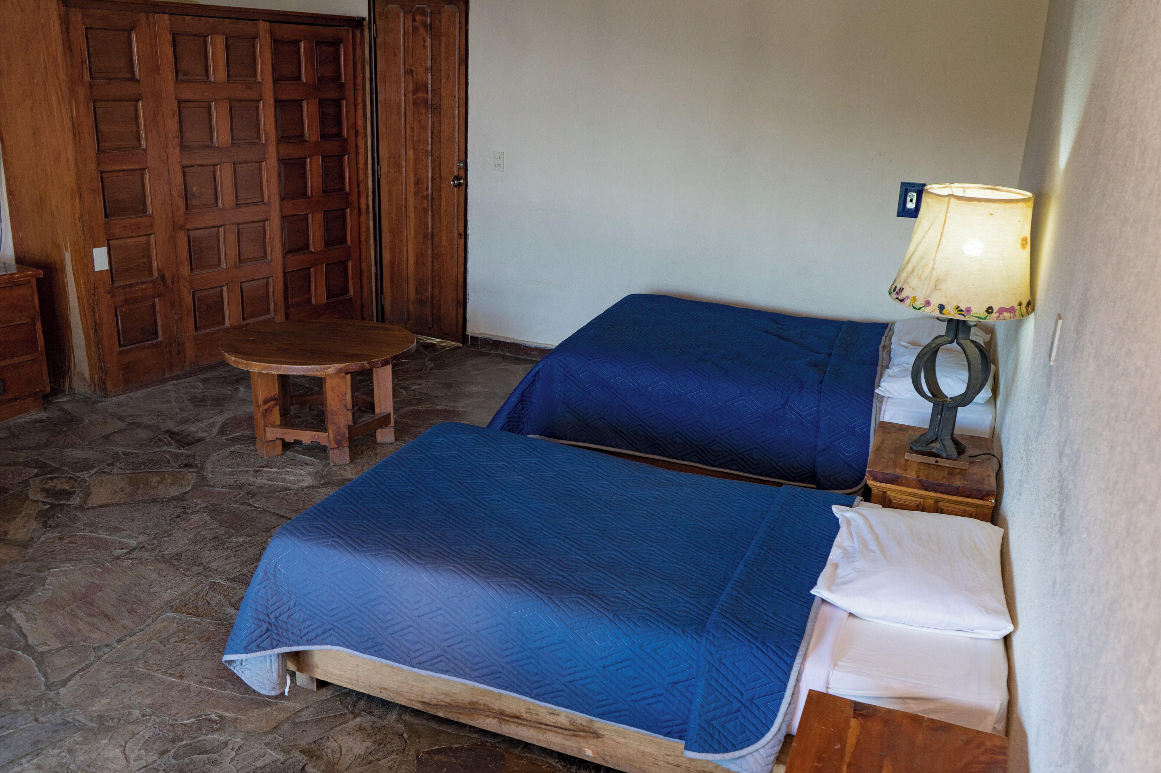 Una habitación con suelo de piedra, dos camas con ropa de cama azul, una puerta de madera y una mesa con una lámpara.