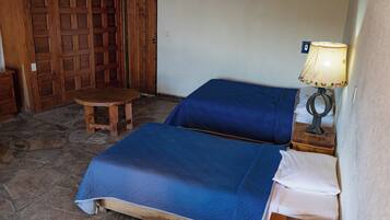 Una habitación con suelo de piedra, dos camas con ropa de cama azul, una puerta de madera y una mesa con una lámpara.