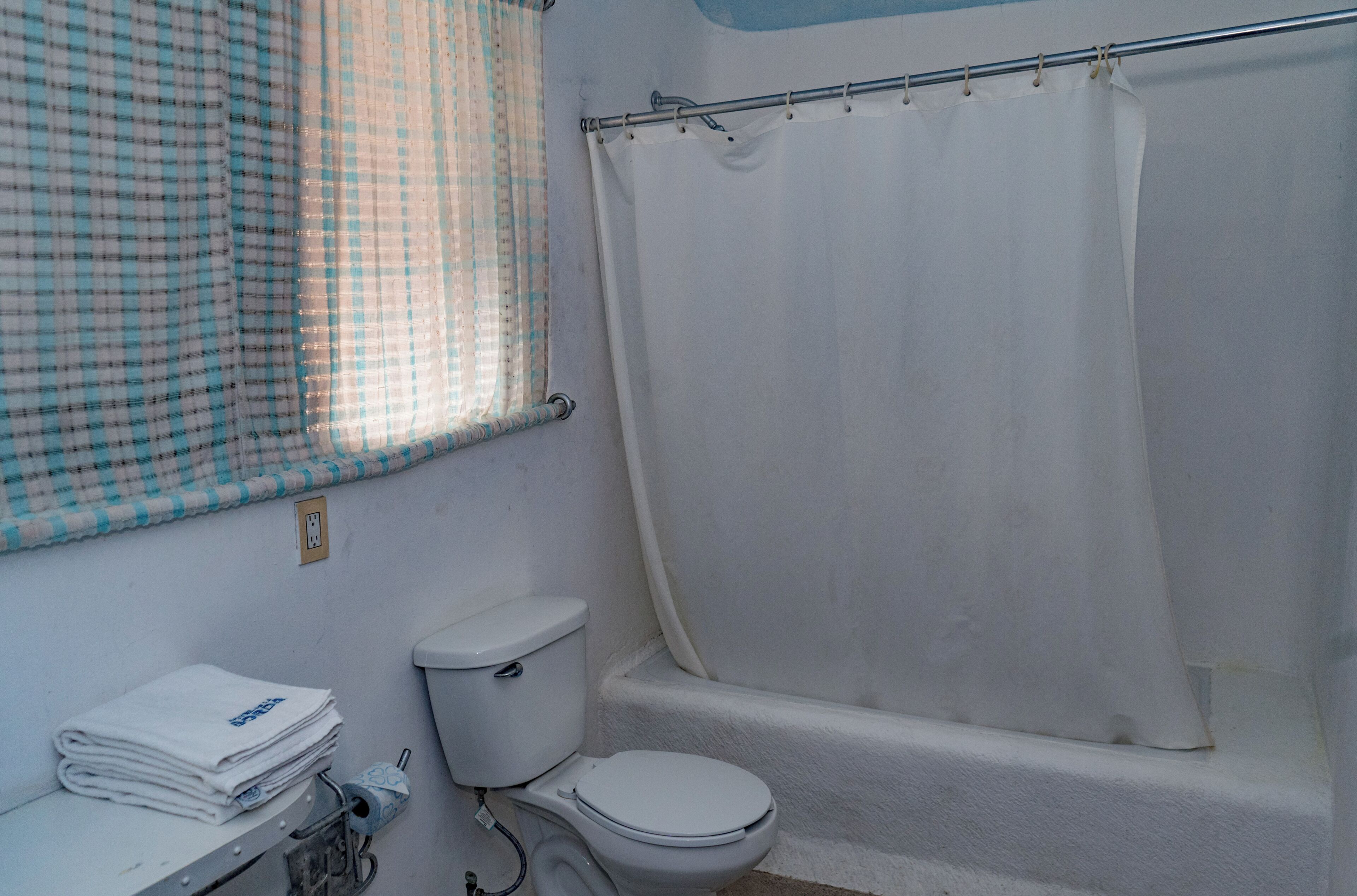 Un baño con inodoro, bañera, ventana con cortina a cuadros y un perchero con toallas.