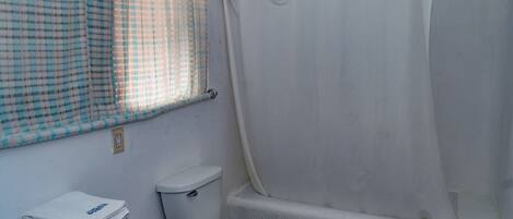 Un baño con inodoro, bañera, ventana con cortina a cuadros y un perchero con toallas.