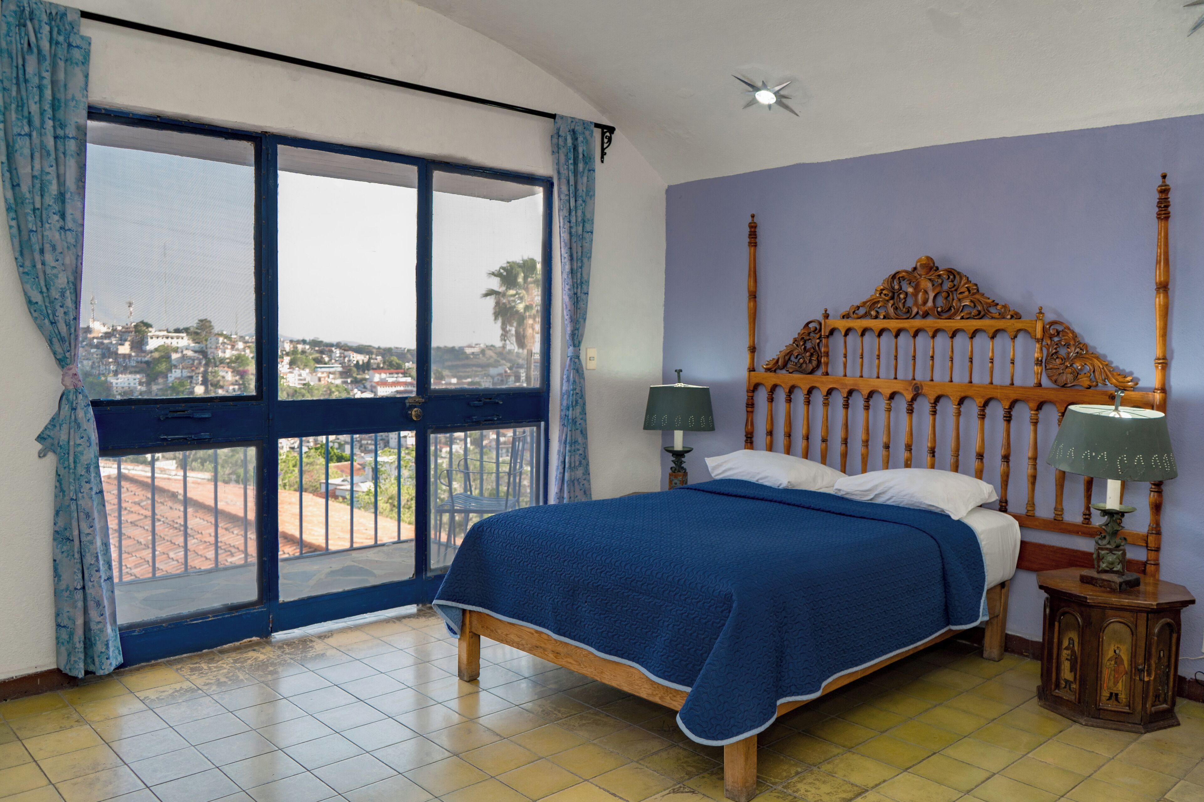 A bedroom with a large window, a bed with a wooden headboard, a nightstand with a lamp, and a view of a cityscape.