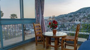Un balcón con una mesa y sillas de madera, un jarrón con flores y vistas a un paisaje urbano.