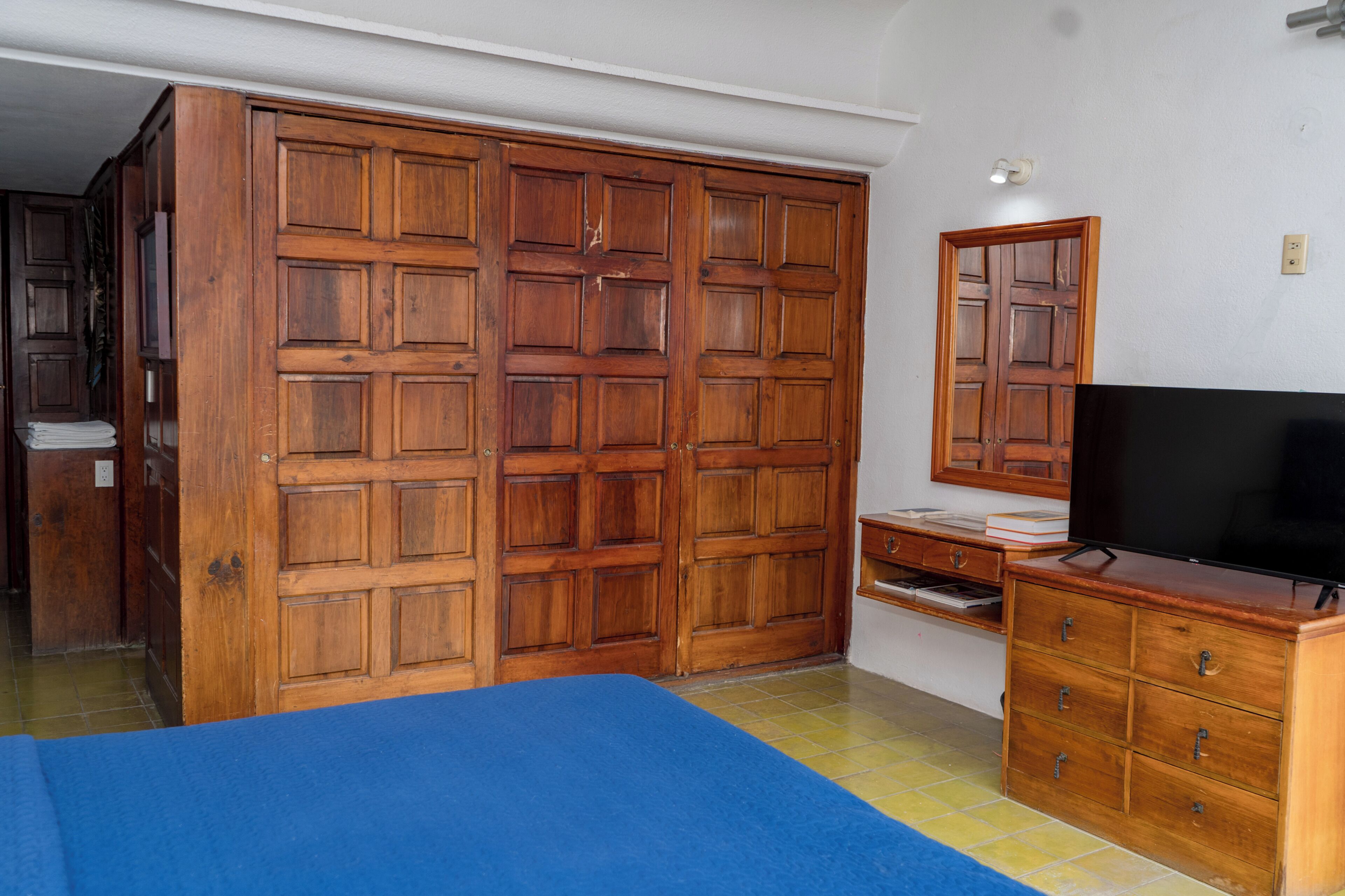 A bedroom with a wooden wardrobe, a television on a wooden dresser, and a bed with a blue bedspread.