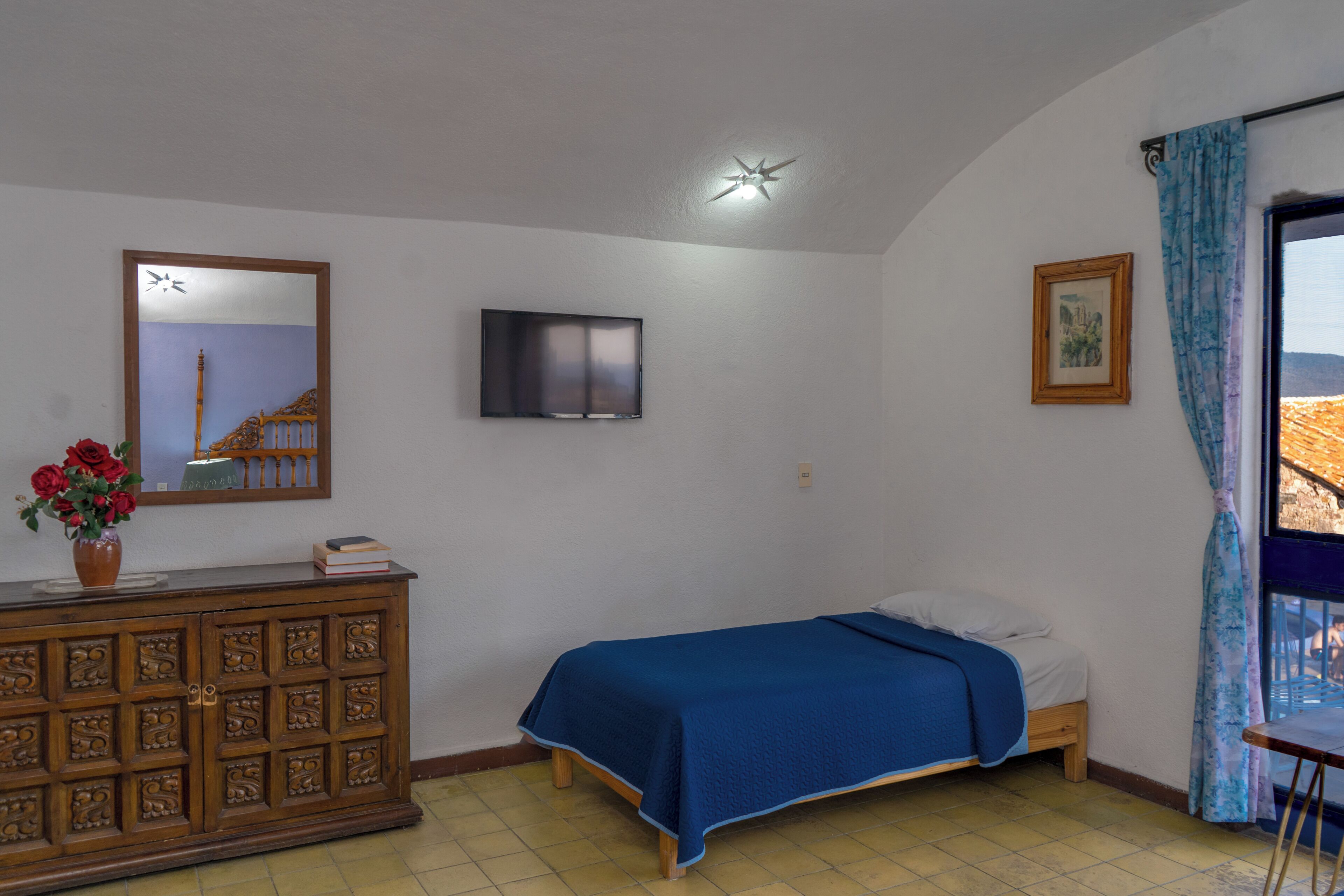 A bedroom with a bed, a dresser, a television, a window with curtains, and a painting on the wall.