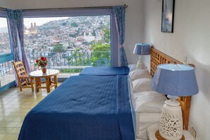 Un dormitorio con cama, una mesita de noche con lámpara, una pequeña mesa con un florero y vistas a un paisaje urbano a través de un balcón.