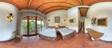 A spacious room with a large bed, a smaller bed, and a bench. There is a balcony with a view of greenery.