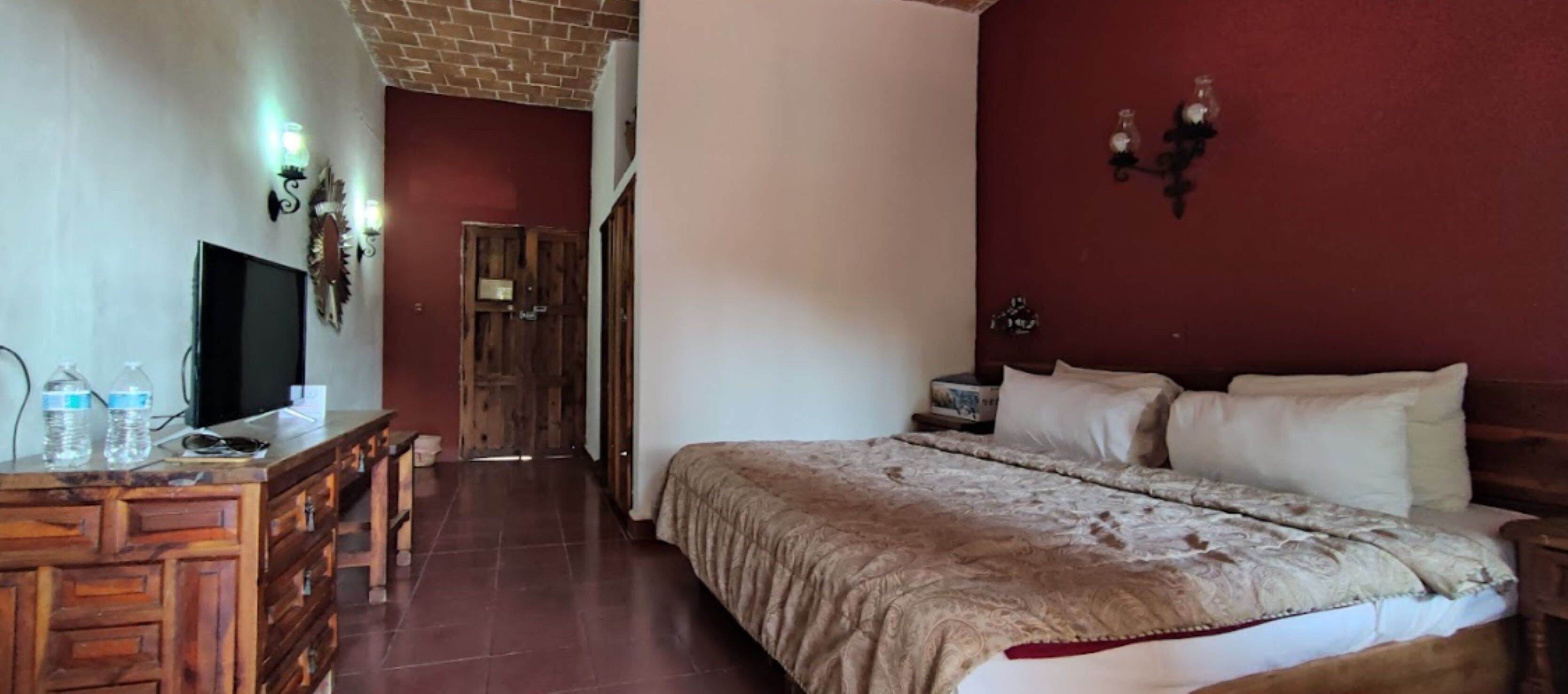 A bedroom with a bed, a wooden dresser, a television, and a wall-mounted light fixture.