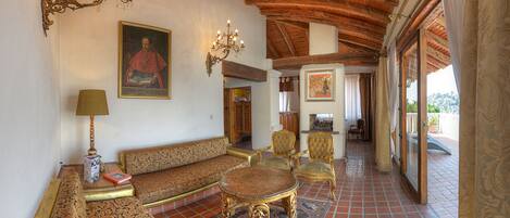 A traditional room with a patterned sofa, a round table, and ornate chairs. There is a chandelier and a painting on the wall.