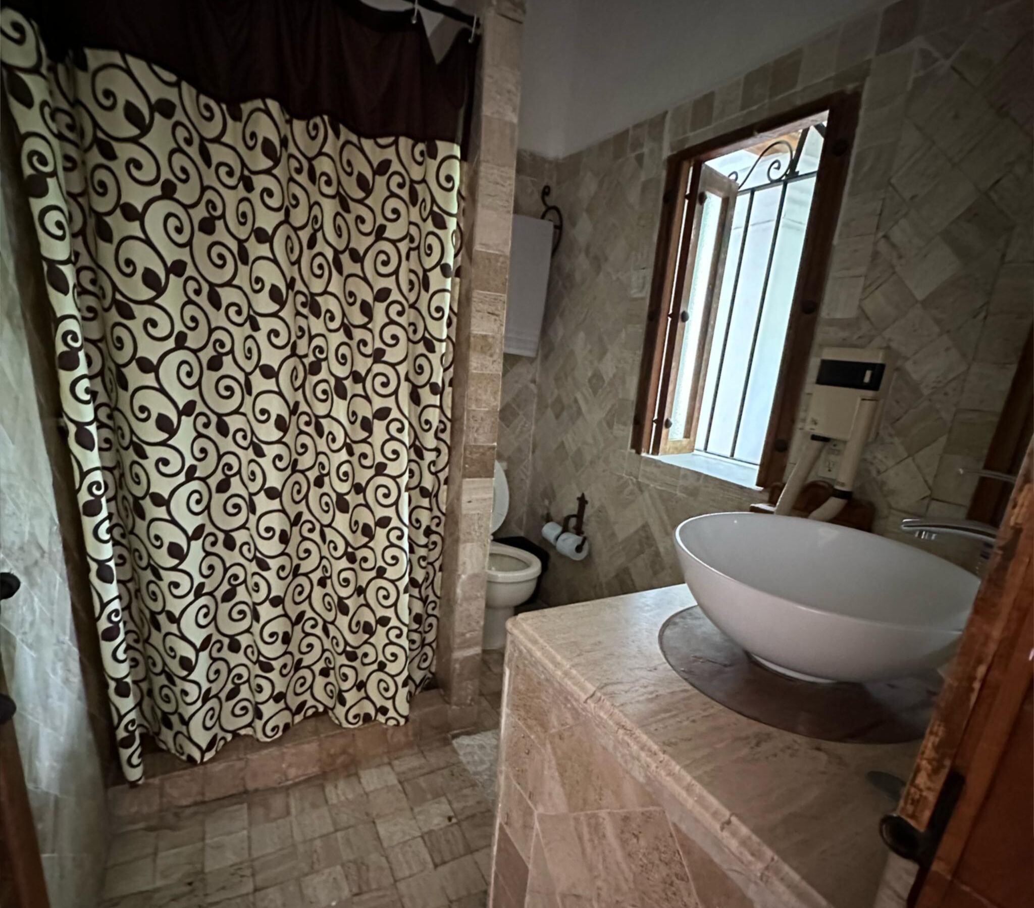 Een badkamer met een gordijn met een patroon, een toilet, een wastafel met een groot bassin en een raam met tralies.