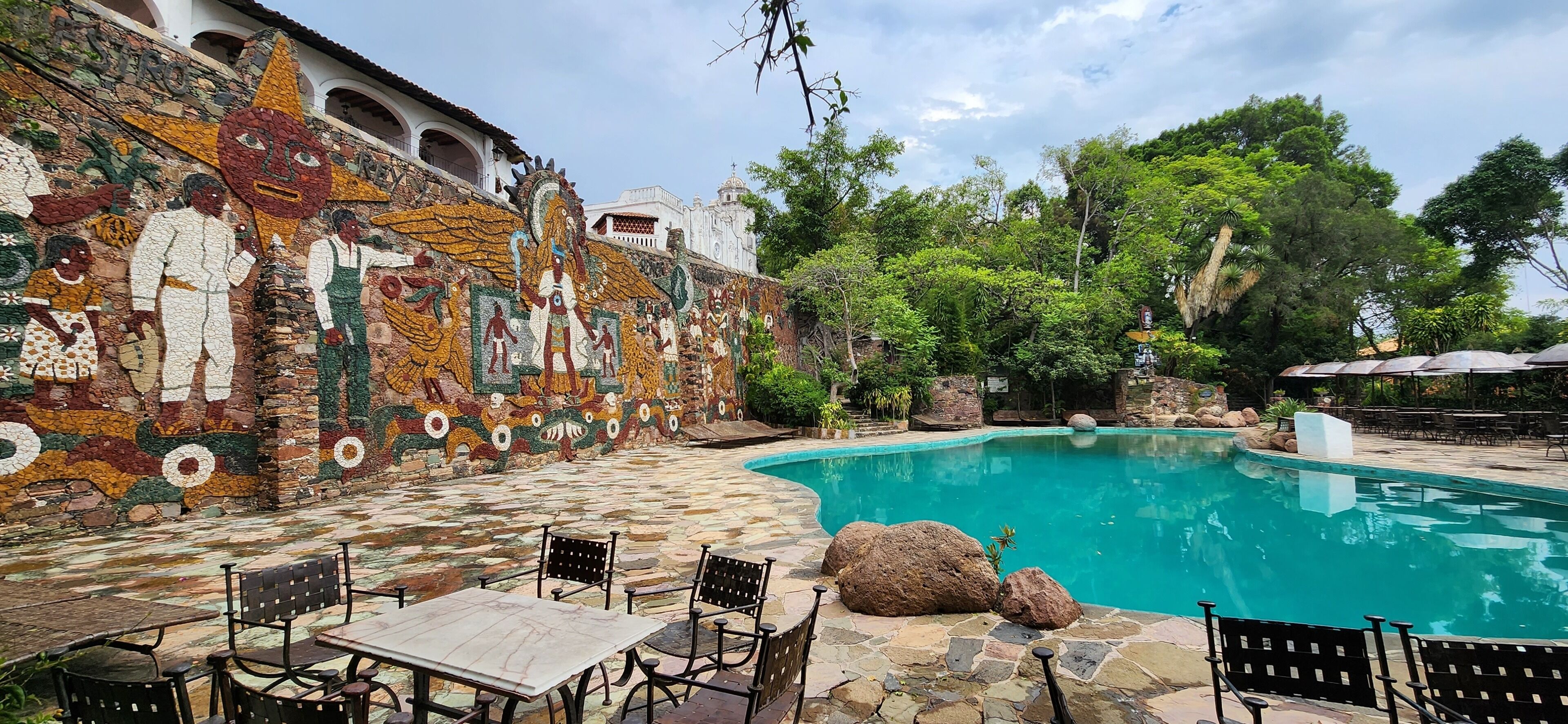 A pool area with mosaic artwork on the walls, outdoor seating, and a clear blue pool.