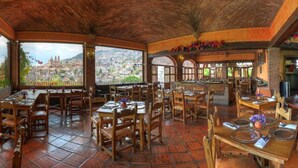 Un restaurante con mesas y sillas de madera, piso de baldosas y vistas a un paisaje urbano a través de amplias ventanas.