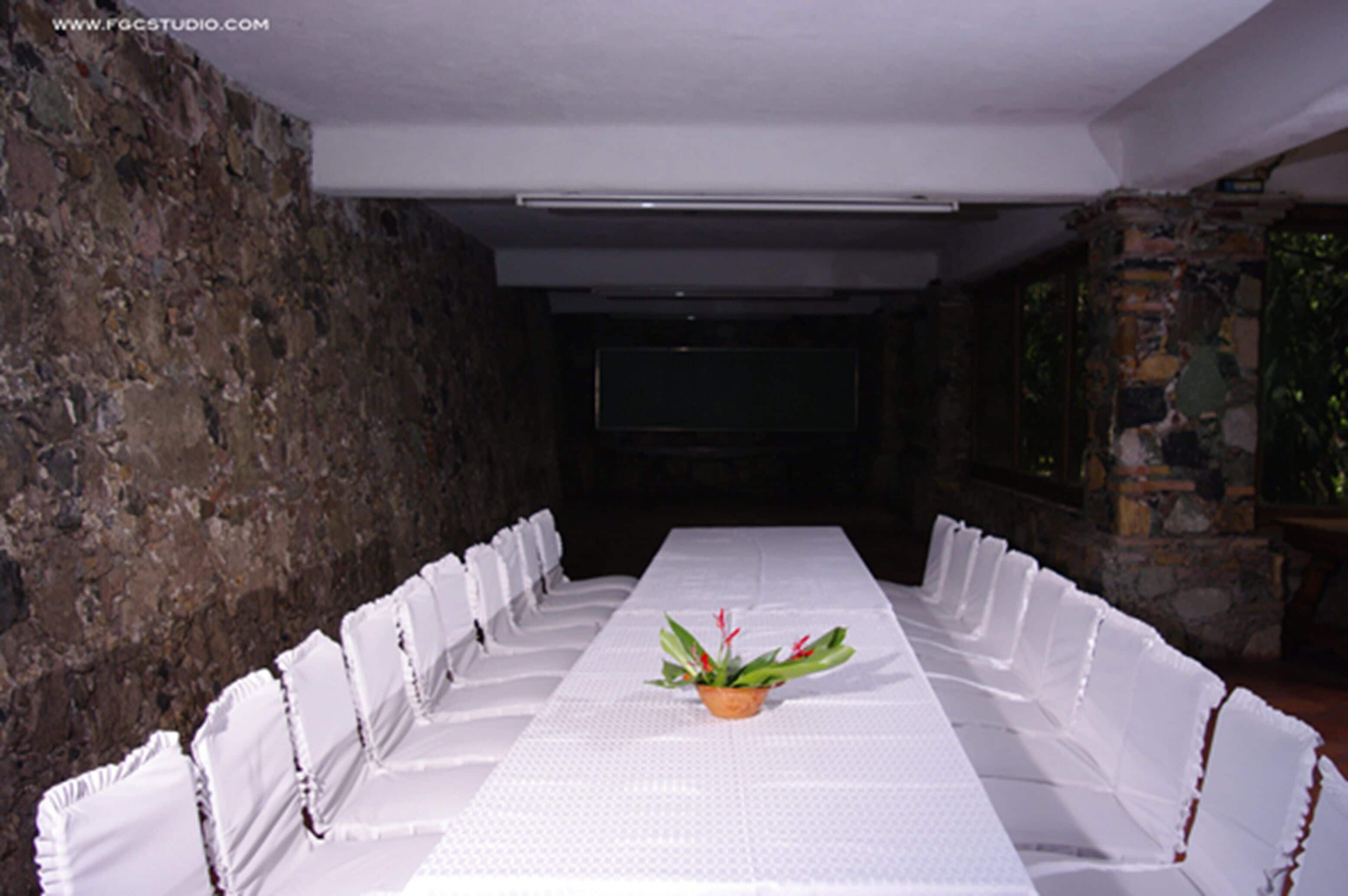 Une longue table à manger, recouverte d’une nappe blanche, entourée de chaises, le tout placé devant un mur de pierre, avec un téléviseur en arrière-plan.