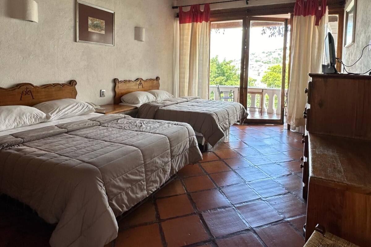 Ein Zimmer mit zwei Betten, einem Holzkasten und einem Balkon mit Vorhängen.