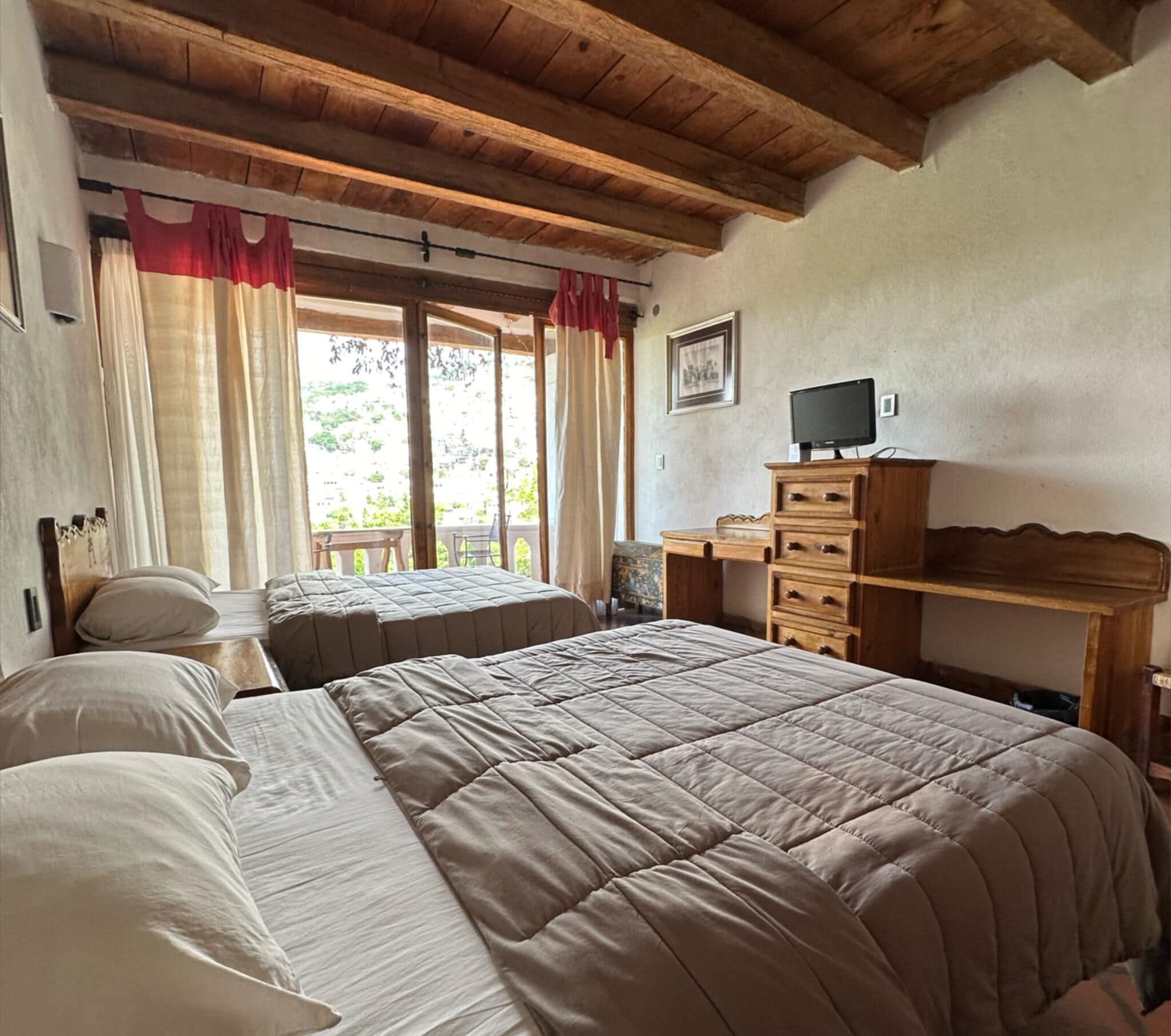 A bedroom with two beds, wooden ceiling beams, a desk with a computer, and a window with curtains.