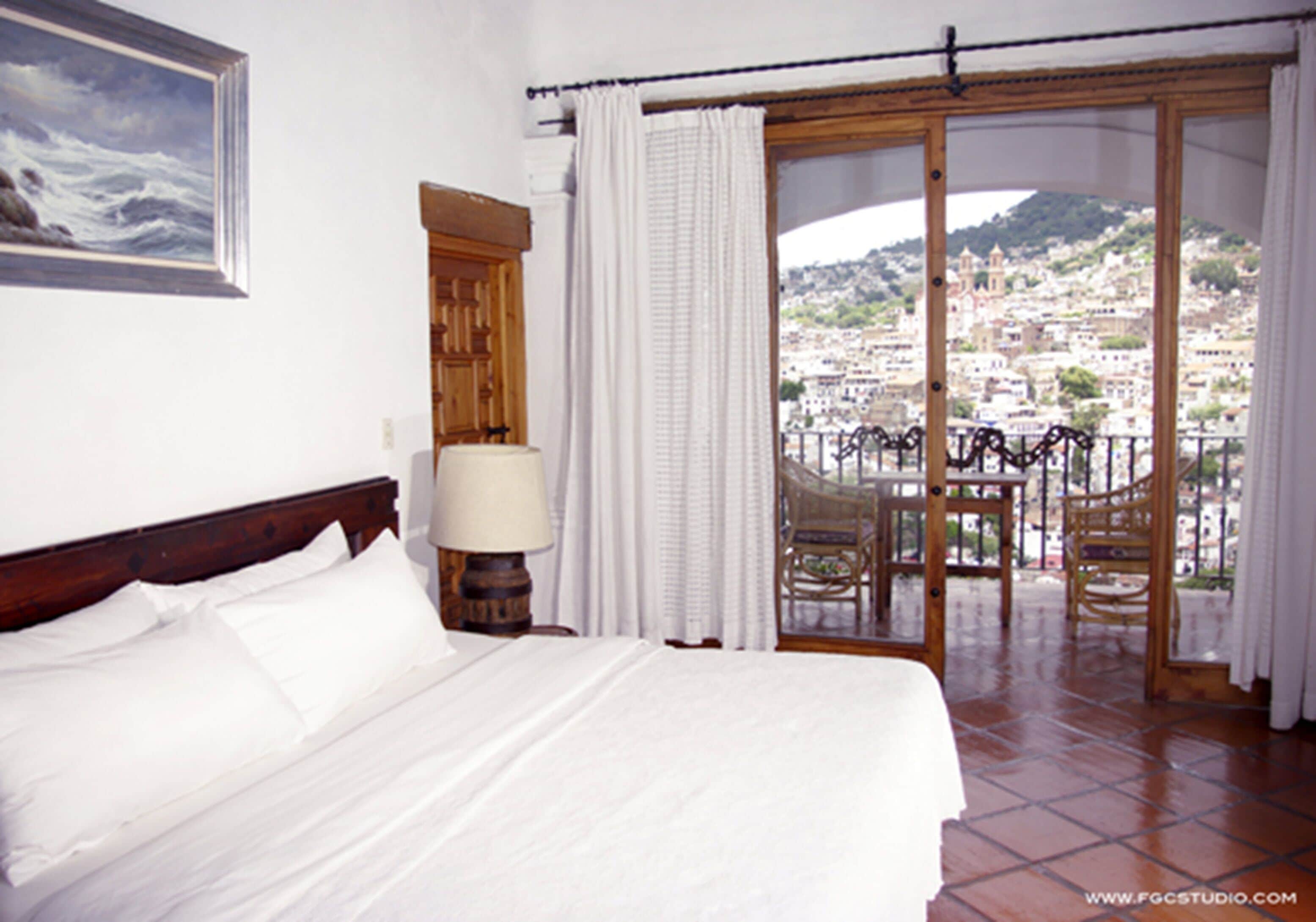 Habitación de hotel con cama, mesita de noche, lámpara, vista a una ciudad a través de una puerta de vidrio y un balcón con sillas.
