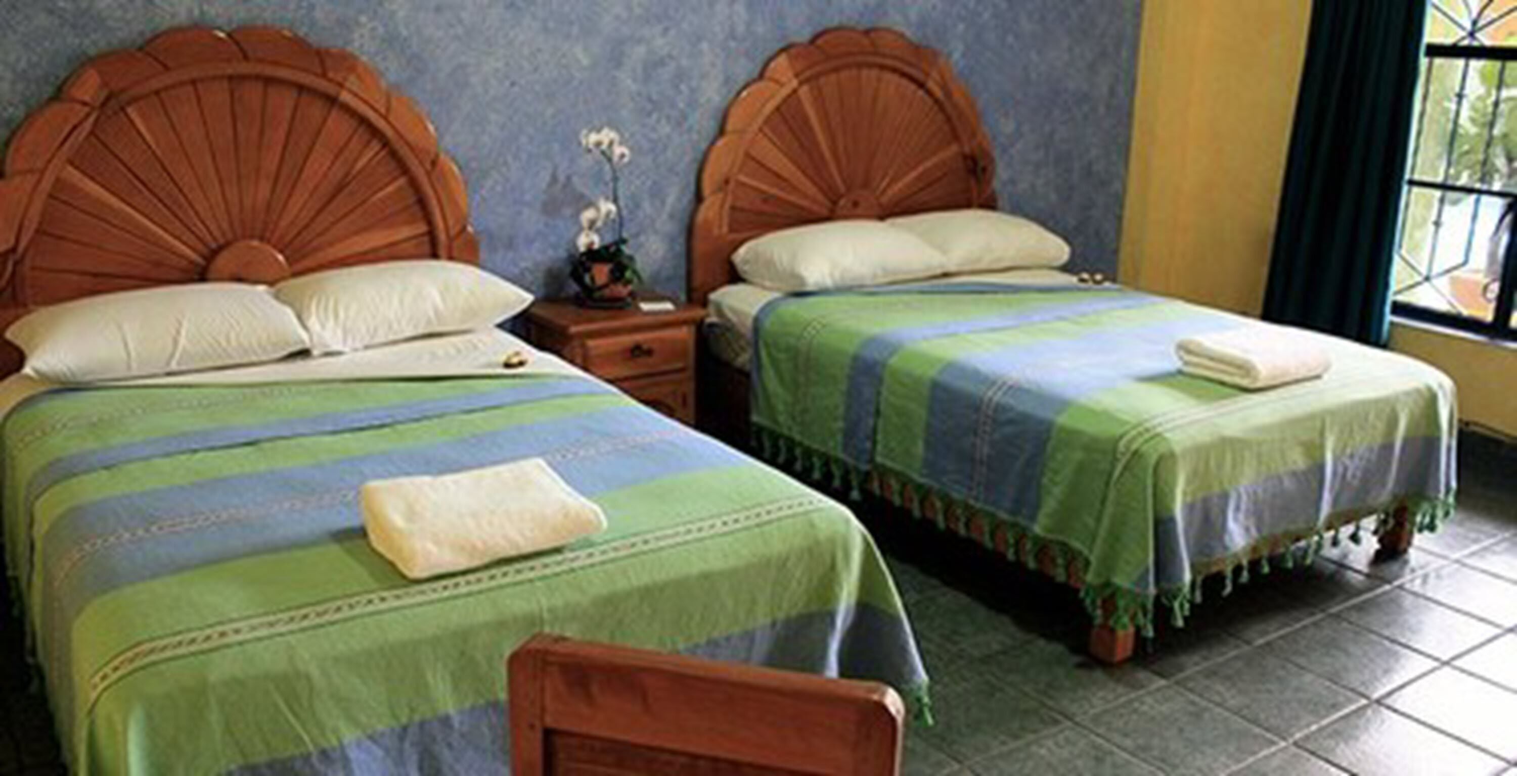 Two single beds with wooden headboards, a nightstand with a small table and an orchid, and a window with curtains.