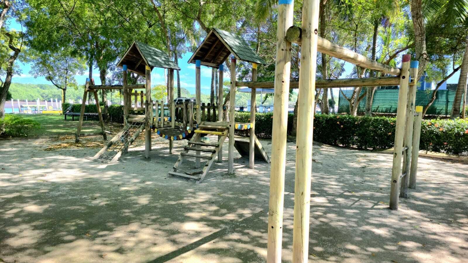 A playground with wooden structures and slides, surrounded by trees and a clear sky.