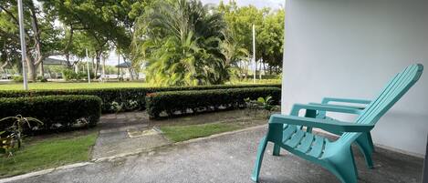 Un espacio exterior techado con una silla turquesa, un sendero pavimentado y vistas a un jardín cuidado con varios árboles y arbustos.