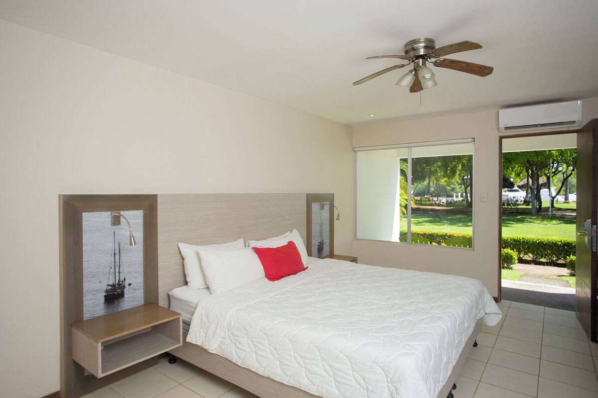 Un dormitorio con cama, mesita de noche, ventilador de techo, ventana con vista a la vegetación y un equipo de aire acondicionado.