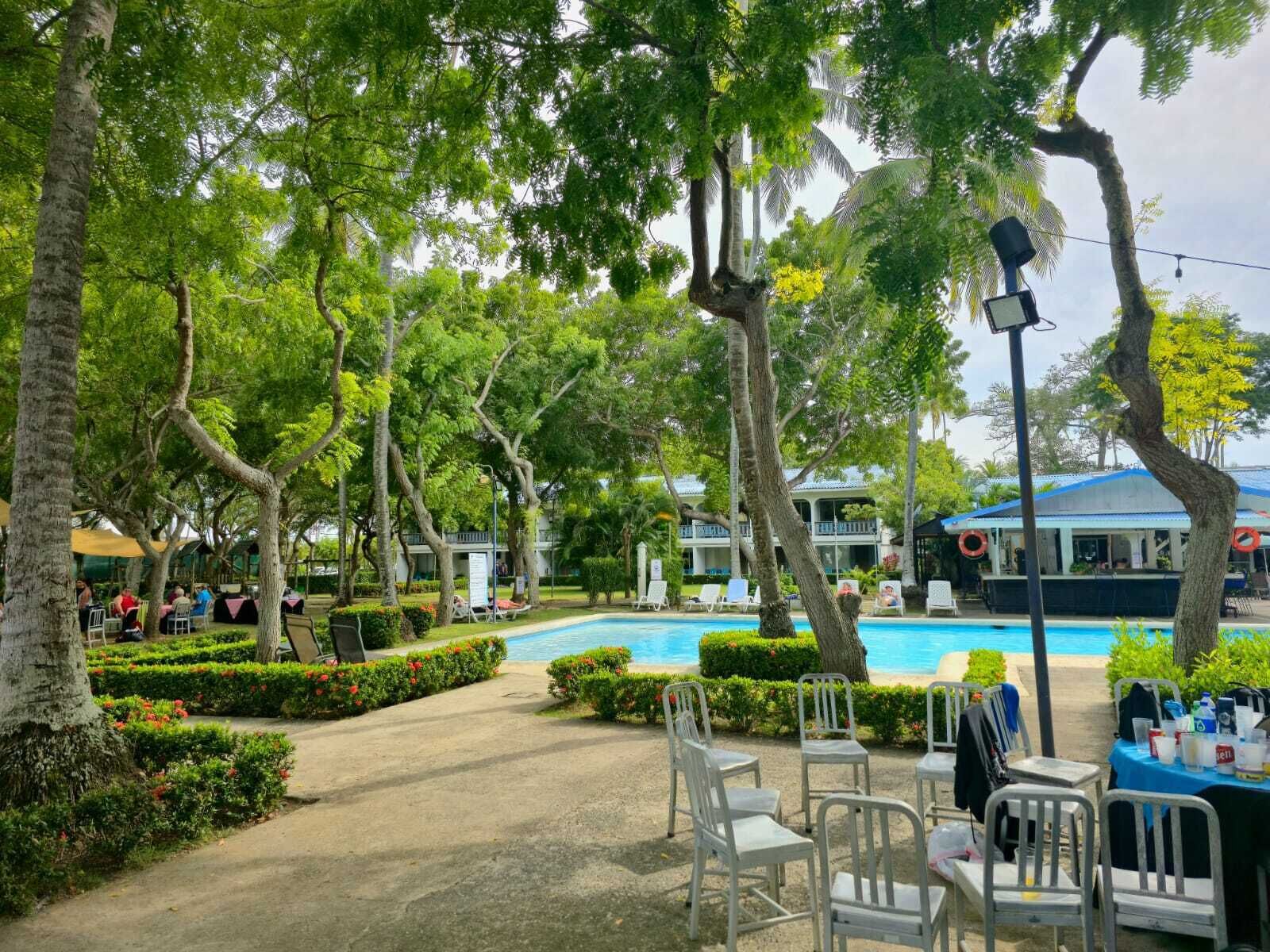 A poolside area with a clear view of the pool, surrounded by trees and a building with a blue roof.