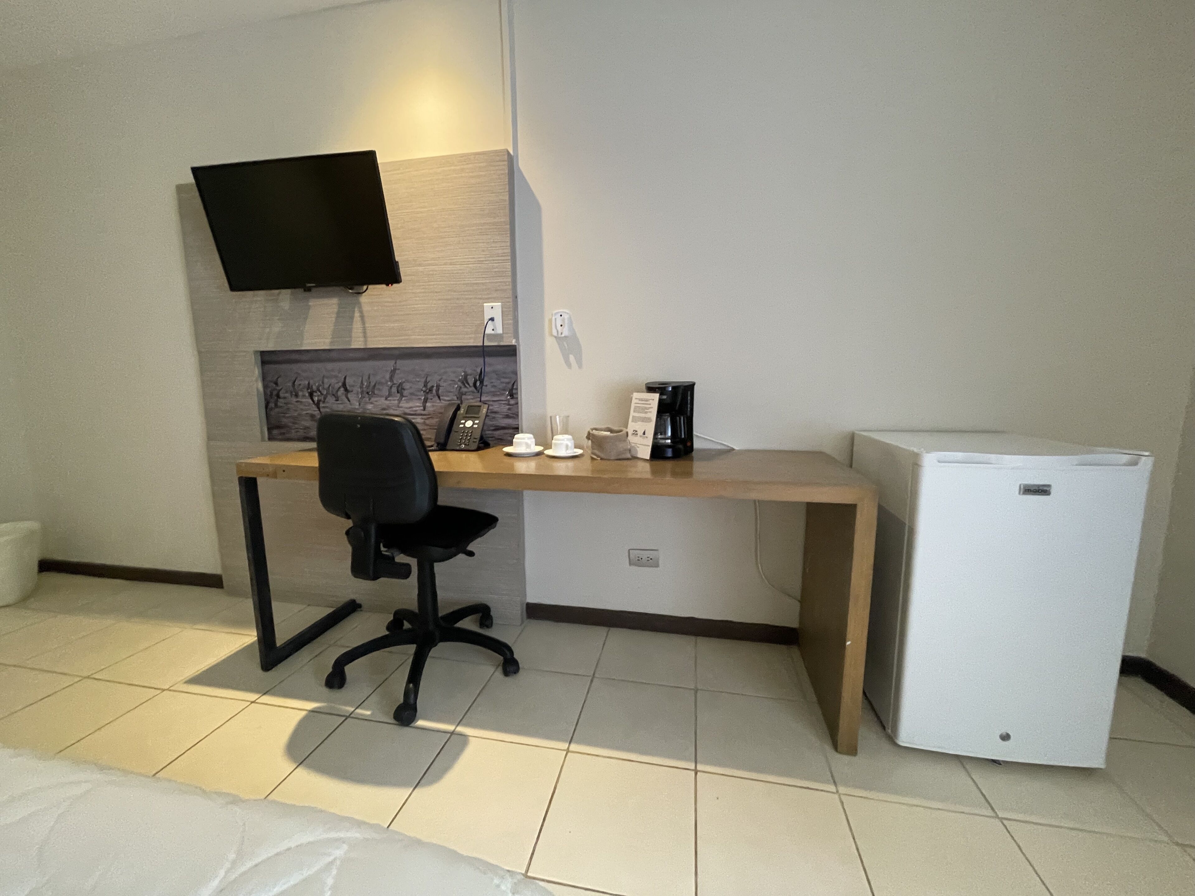 Habitación de hotel con cama, escritorio con silla, televisor montado en la pared, cafetera y refrigerador.