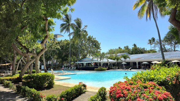 Zona de piscina en un resort con sillones de descanso, un espacio de estar cubierto y exuberante vegetación.