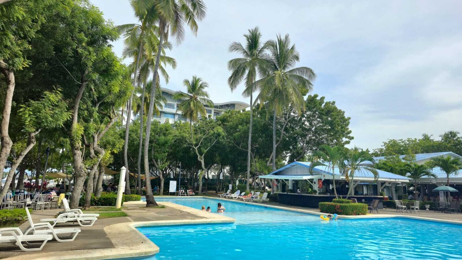 A swimming pool surrounded by palm trees and lounge chairs, with a building in the background.