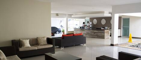 A modern lobby with a sofa, coffee table, and wicker chairs. There is a reception desk with a counter and a car visible through the window.