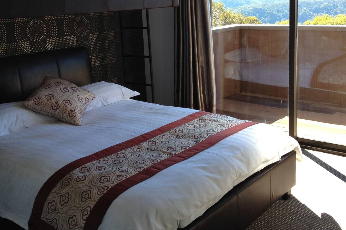 A hotel room with a large bed, a headboard with a patterned design, a window with a view of the outdoors, and a balcony.